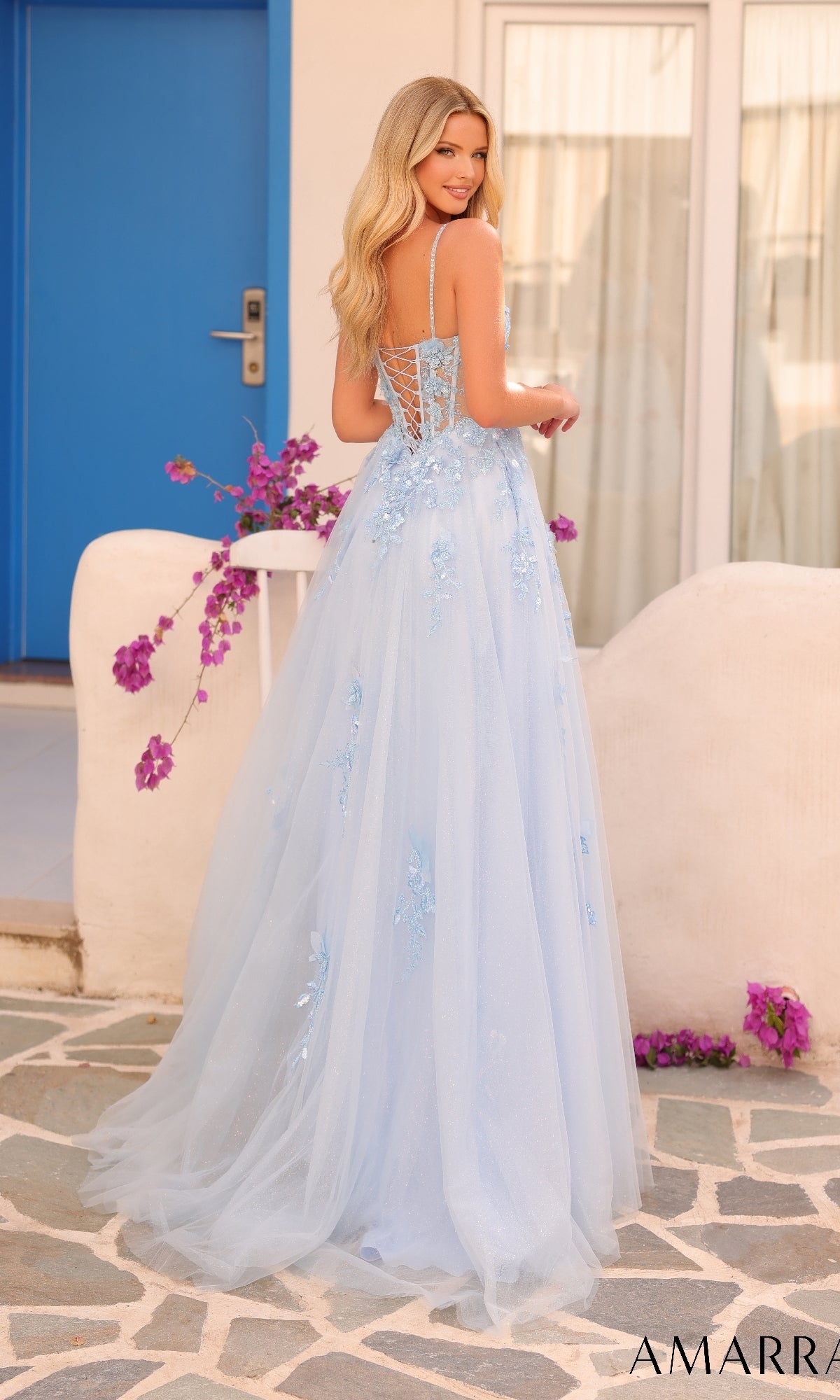 A woman stands outdoors in front of a white building, wearing the Amarra 88086 long formal dress?a light blue, floor-length gown featuring lace embroidery and a delicate lace-up back.